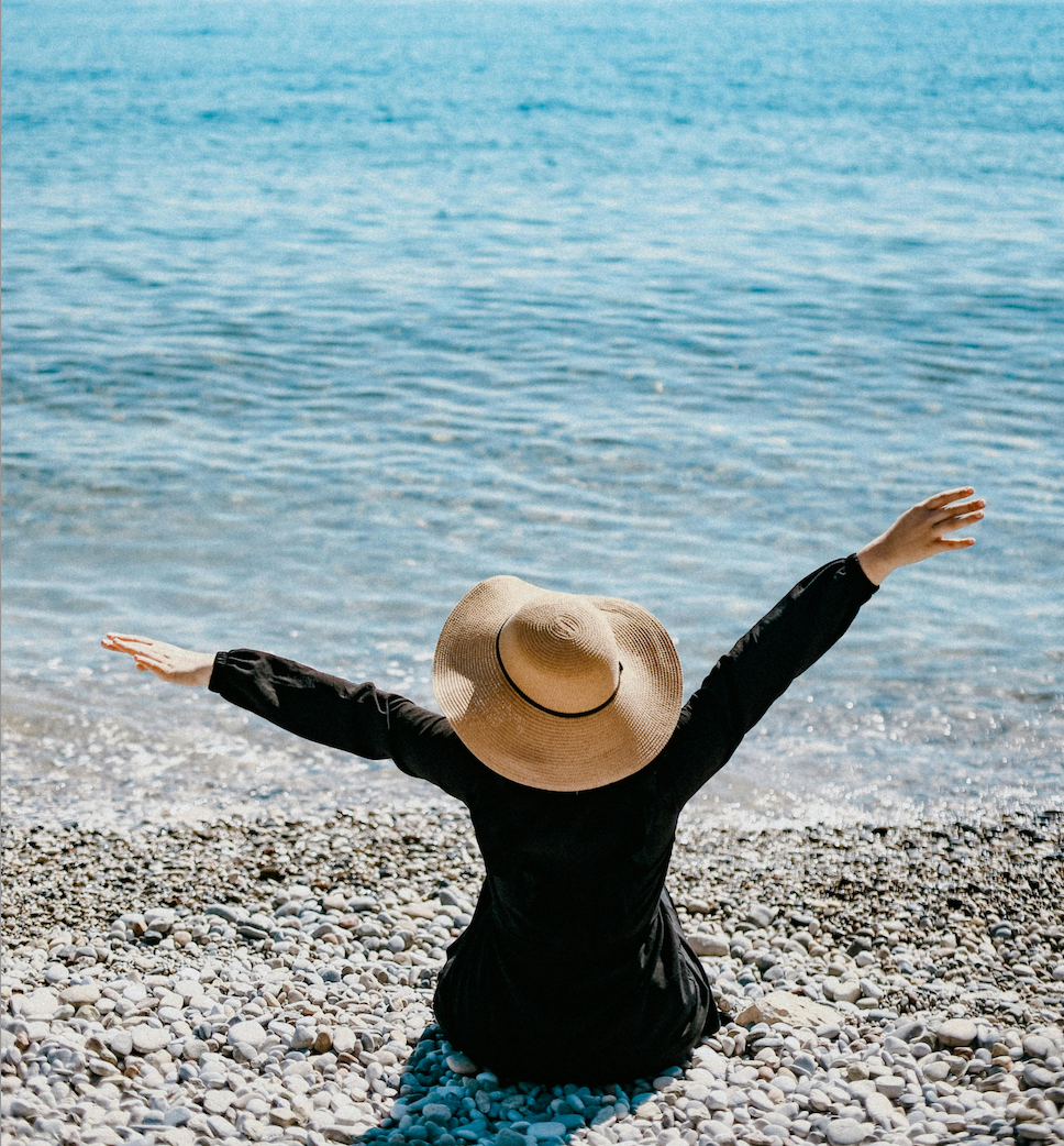 Une femme est fasse à la mer avec un chapeau et les bras en l'air