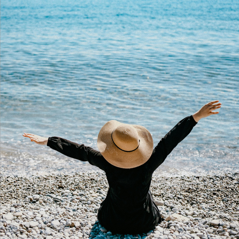 Une femme est fasse à la mer avec un chapeau et les bras en l'air