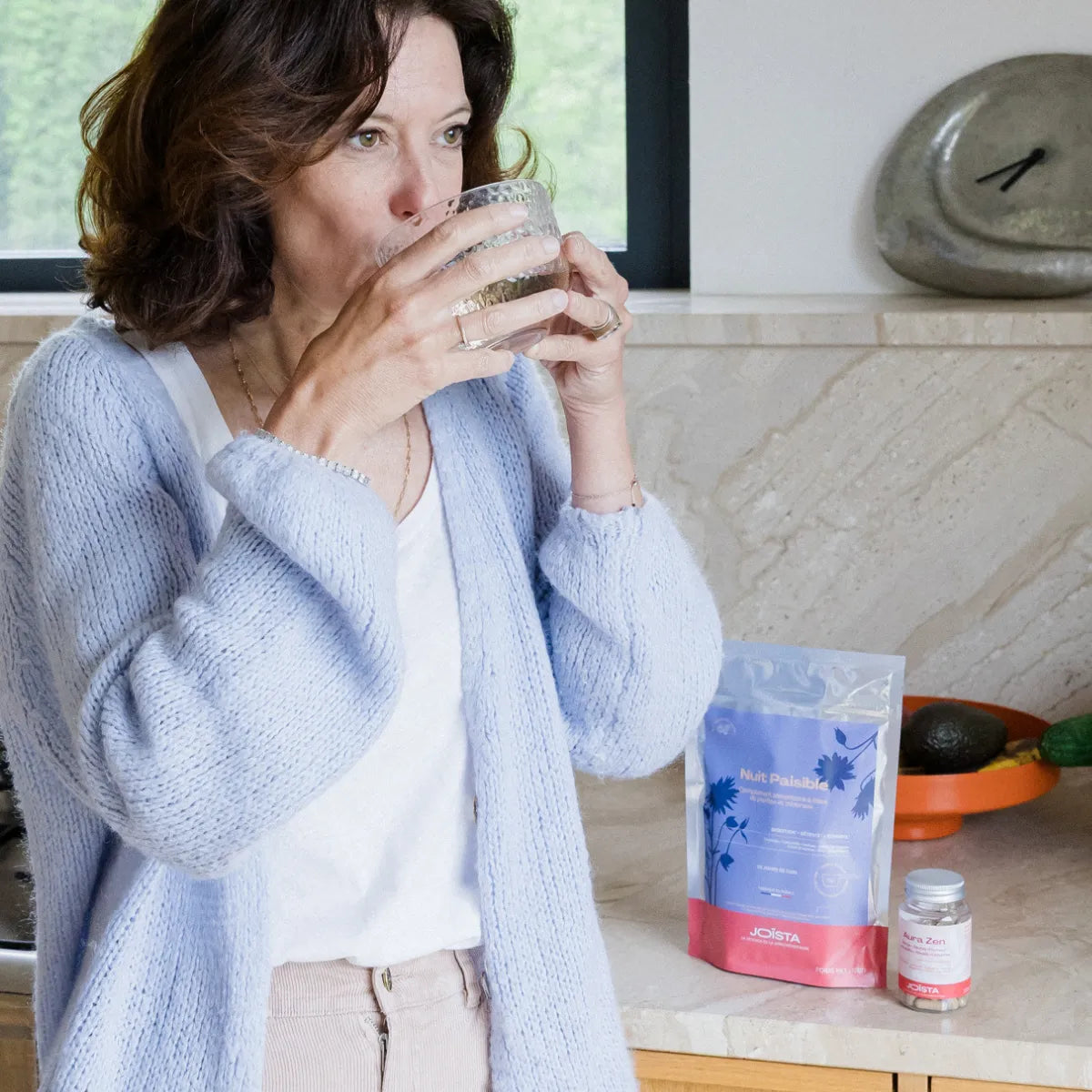 Une femme en périménopause dans sa ciusine boit la boisson calmante et digestive Nuit paisible de Joïsta, dans une tasse, et prend des gélules aura zen de Joïsta.